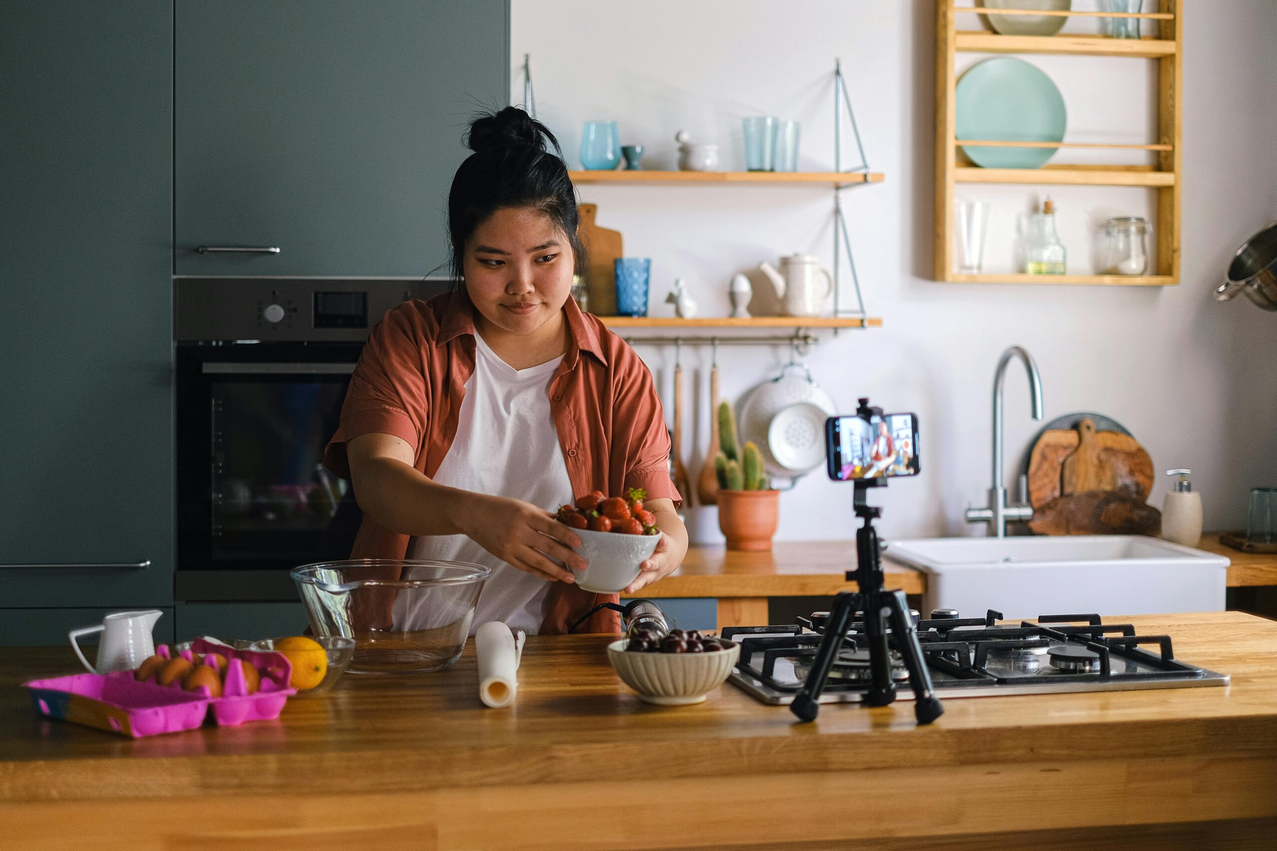 Woman filming a cooking video on a smartphone in a home kitchen setting with fresh ingredients.